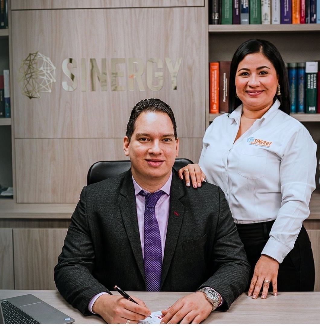 El Dr. Jesús Ramírez en su escritorio con una colaboradora, frente al logo de Sinergy en la pared de la oficina.